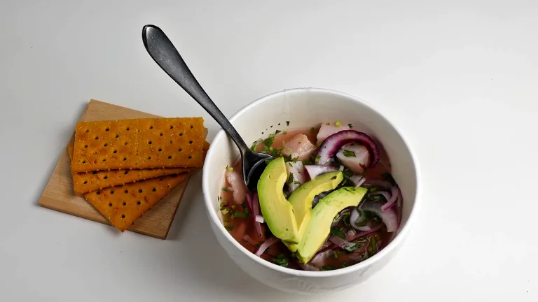 Ceviche de tilapia con cebolla morada, culantro y tajadas de aguacate en un bowl blanco, con galletas al lado