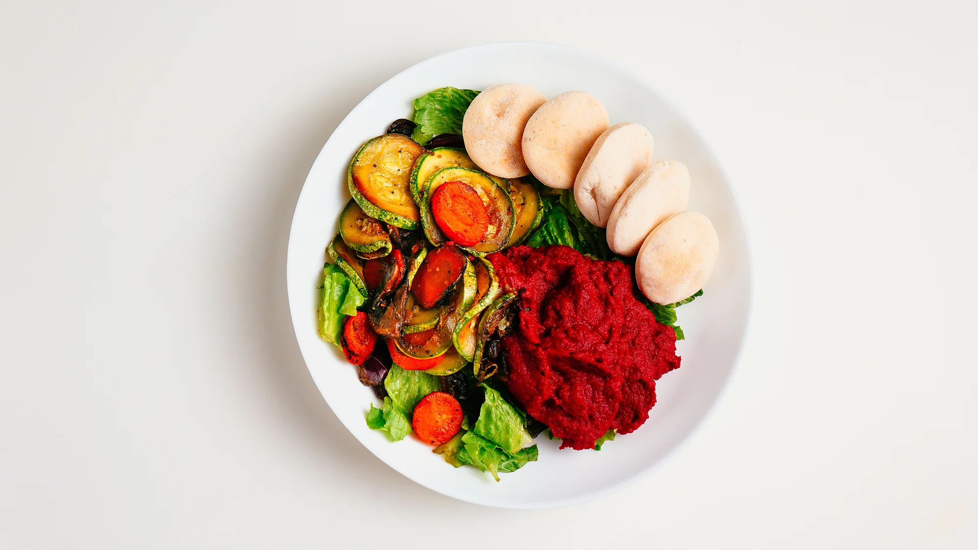 Bowl de hummus de remolacha con vegetales salteados, lechuga y panes pita pequeños, visto desde arriba
