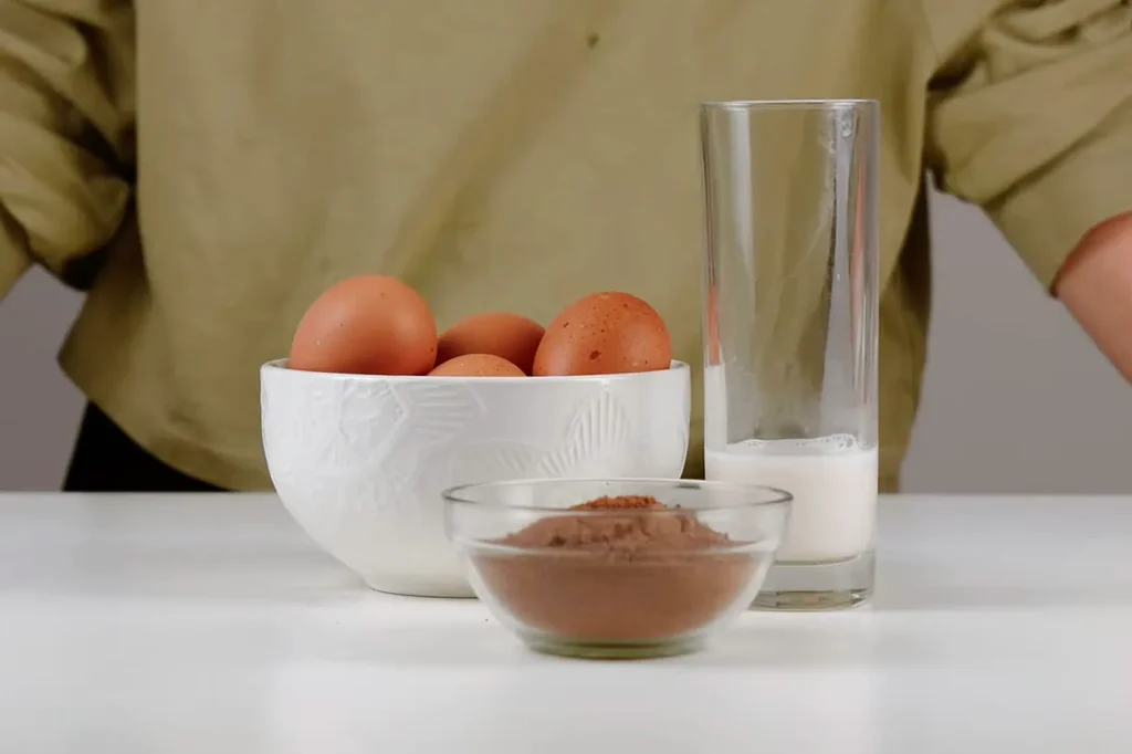Los ingredientes del mousse: un bowl con huevos, un bowl de vidrio con chocolate en polvo y un vaso de leche de almendras