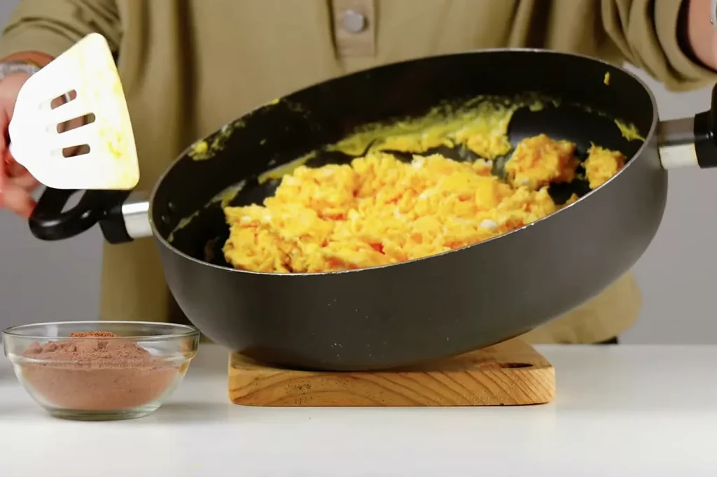 Huevos revueltos bien cocidos en el sartén, sin nada líquido, junto al bowl de chocolate en polvo